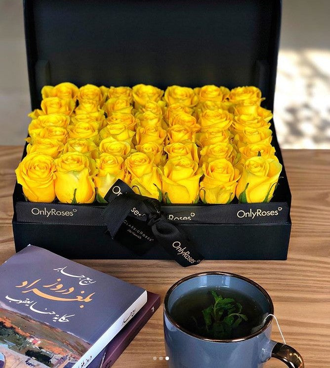 Box of yellow roses with 'OnlyRoses' branding, a book, and a cup of tea on a wooden surface.
