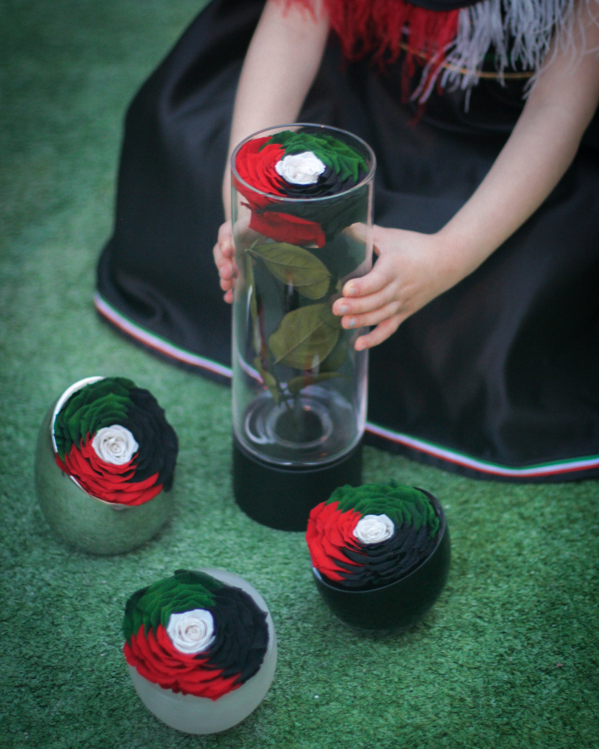 Person holding a cylindrical container with a preserved rose, surrounded by smaller rose arrangements on a green surface.