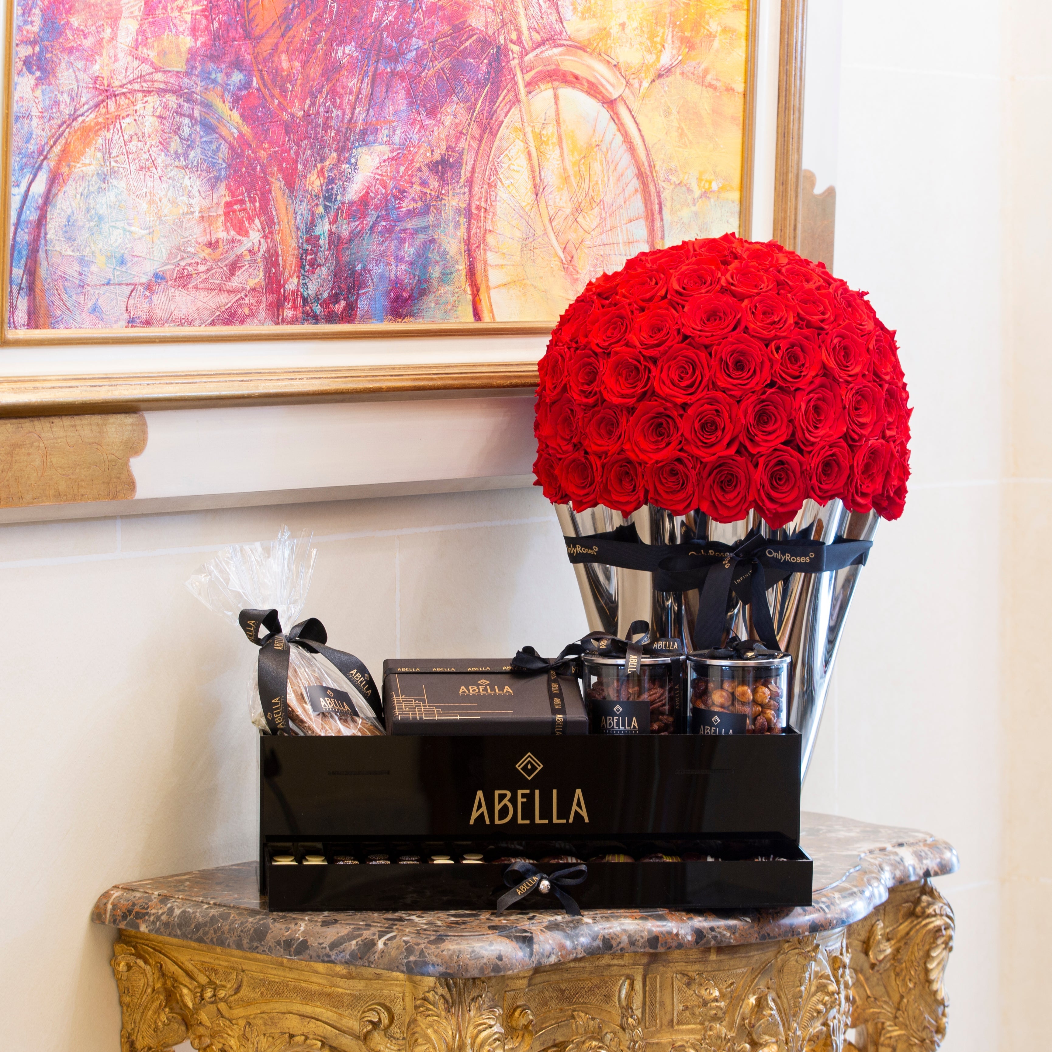 Decorative setup with red roses, Abella products, and a colorful painting in the background.