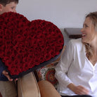 Person holding a heart-shaped box of red roses to a smiling woman.